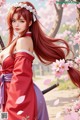 A woman with long red hair in a traditional red and purple outfit stands amidst cherry blossoms.